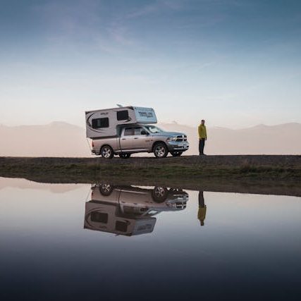 Ein Wohnmobil steht am Rand einer Straße, reflektiert im Wasser, mit einer Person daneben.